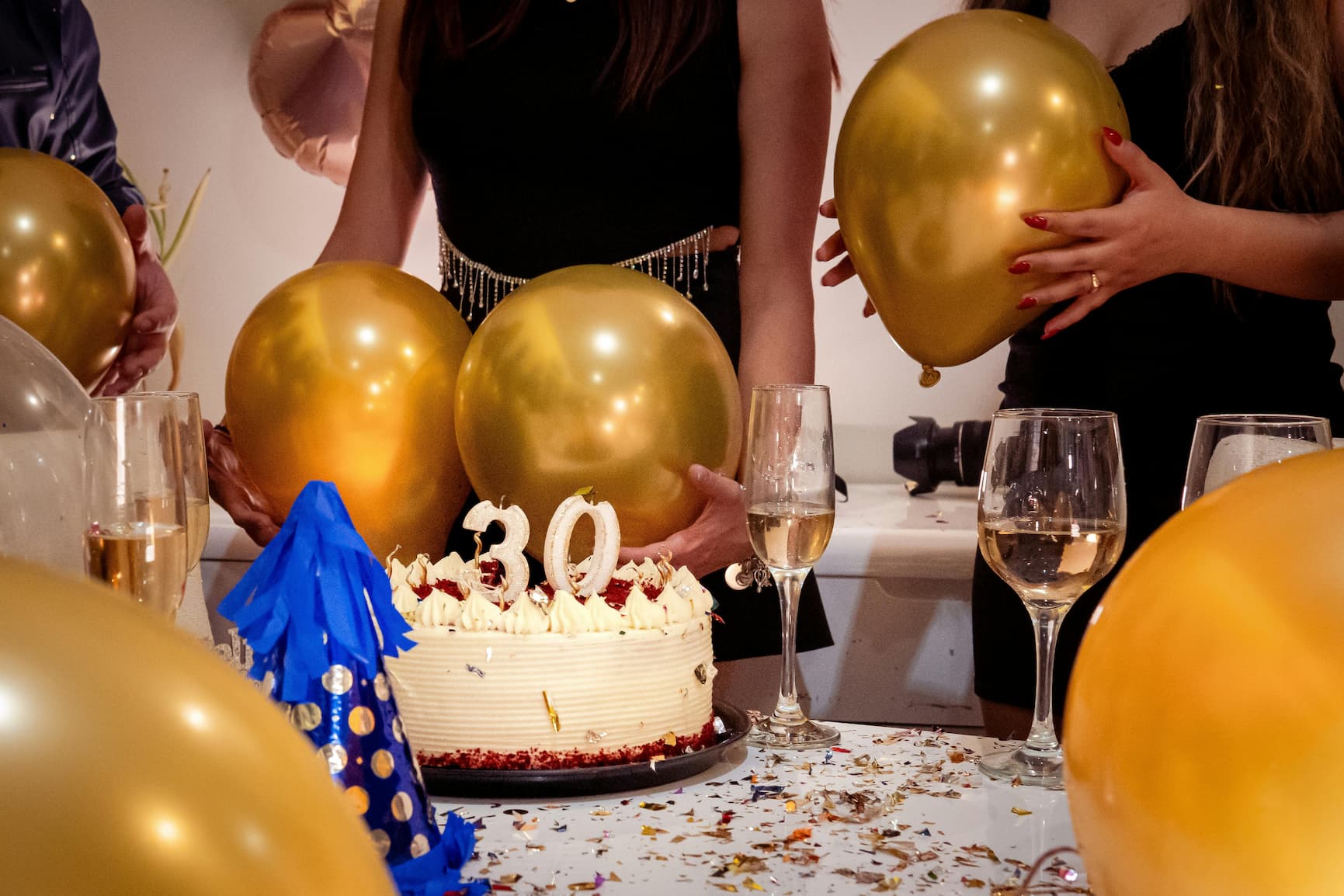 A group of people gathered around a colorful cake, smiling, with balloons floating above them.