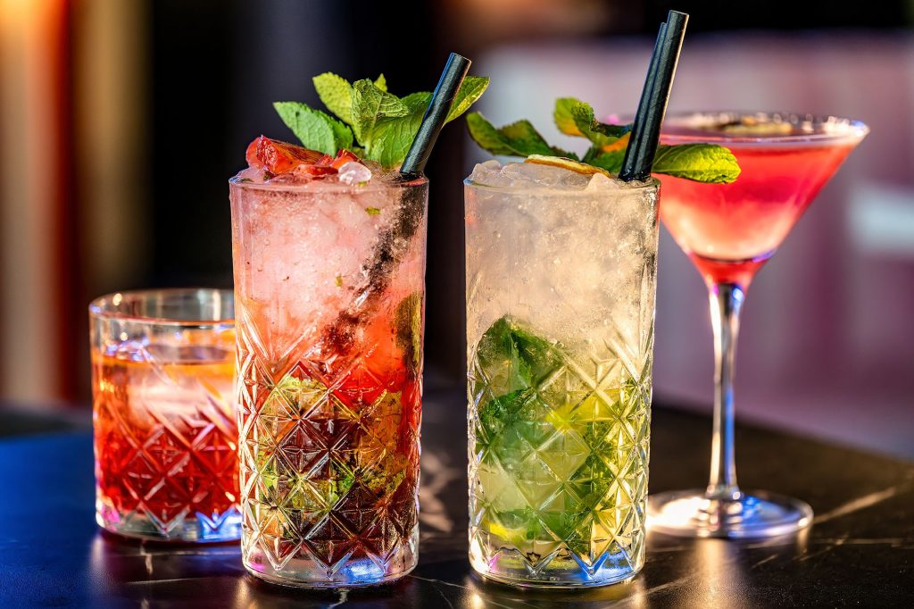 Three colorful cocktails are displayed on a table, showcasing different garnishes and glass styles.