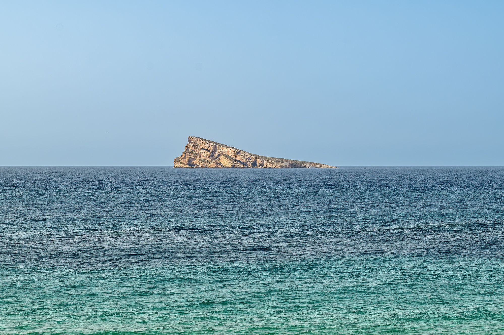 A large rock stands alone in the ocean near Benidorm Beach, surrounded by blue water and a clear sky.