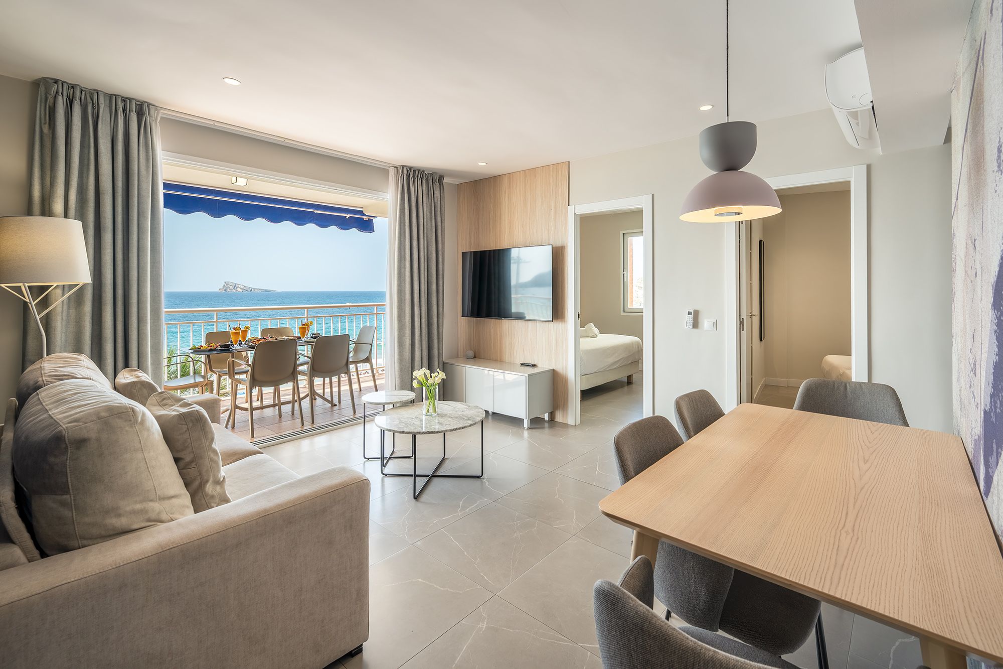 Bright living room in Mara boutique apartment, complete with dining table, TV, and a balcony overlooking the ocean.