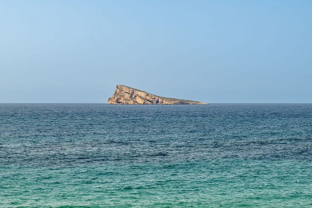 A large rock stands alone in the ocean near Benidorm Beach, surrounded by blue water and a clear sky.