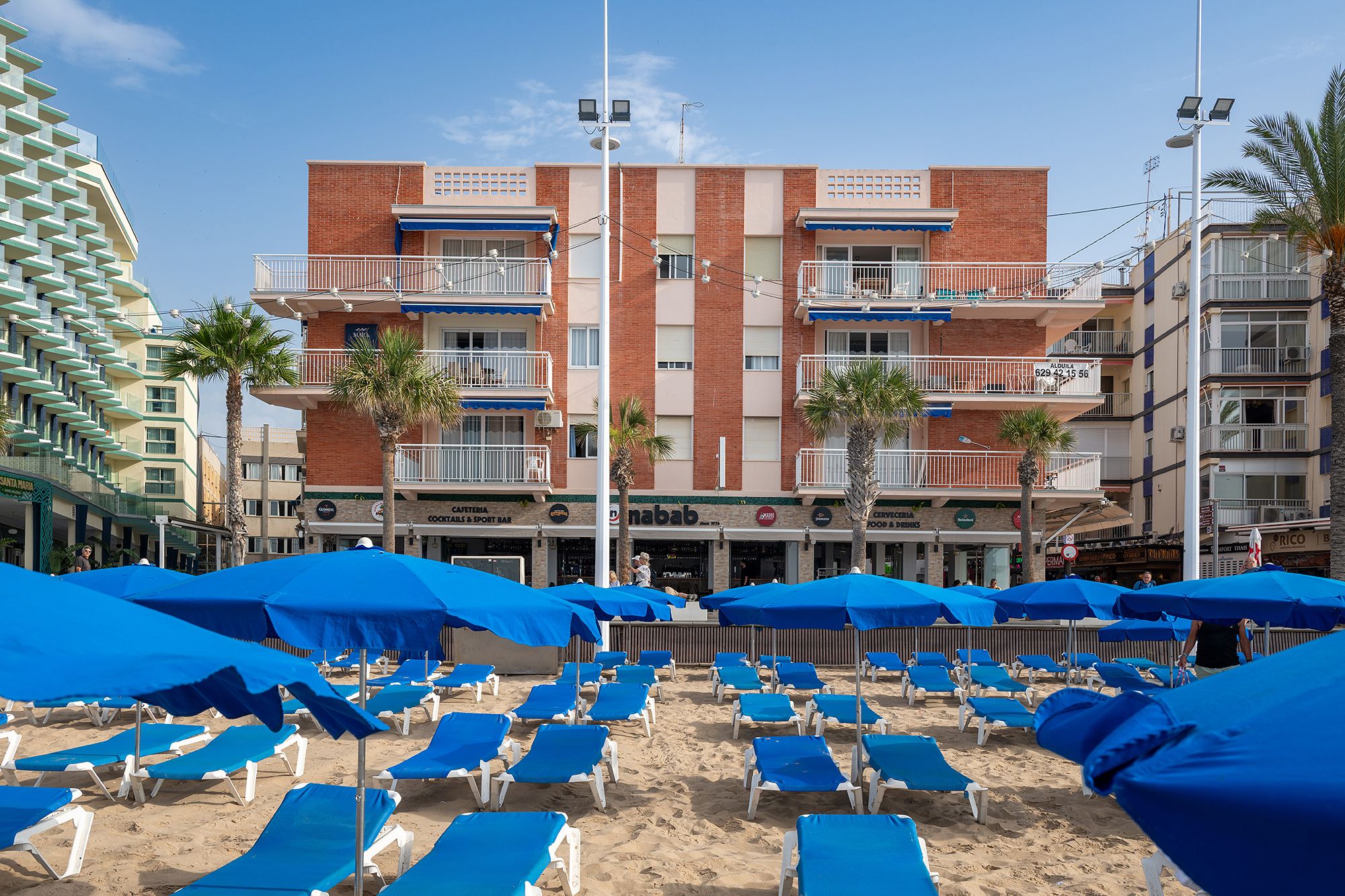 A beach scene featuring blue umbrellas and chairs, with the Benidorm Apartment Mara Boutique in the background.