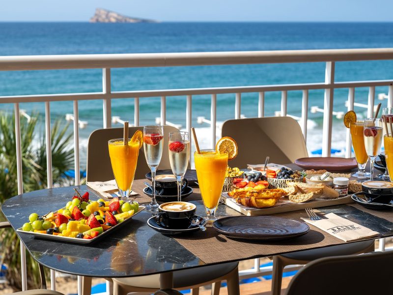 Una pintoresca escena de balcón con una mesa adornada con comida y bebida, con vistas a las serenas olas del océano.